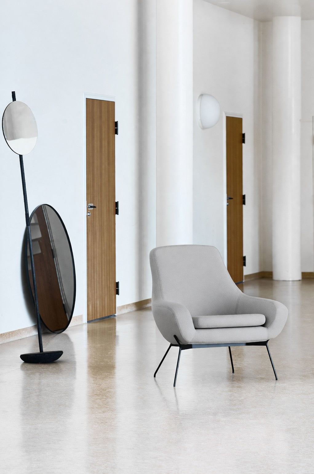 Une pièce moderne et minimaliste avec des murs clairs, deux miroirs ovales sur pied, trois portes en bois et le fauteuil gris Fauteuil Lounge Nomi de Susanne GRØNLUND pour Monsieur Bureau - parfait pour le design contemporain.#finitions_gris_clair