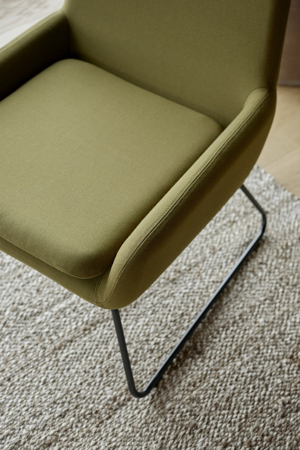 Gros plan sur le Fauteuil Luge Boreale - Design Busk+Hertzog by Monsieur Bureau, composé d'un tissu vert olive et d'une base luge en acier noir, posé sur un tapis tissé beige texturé sur un sol en bois.
