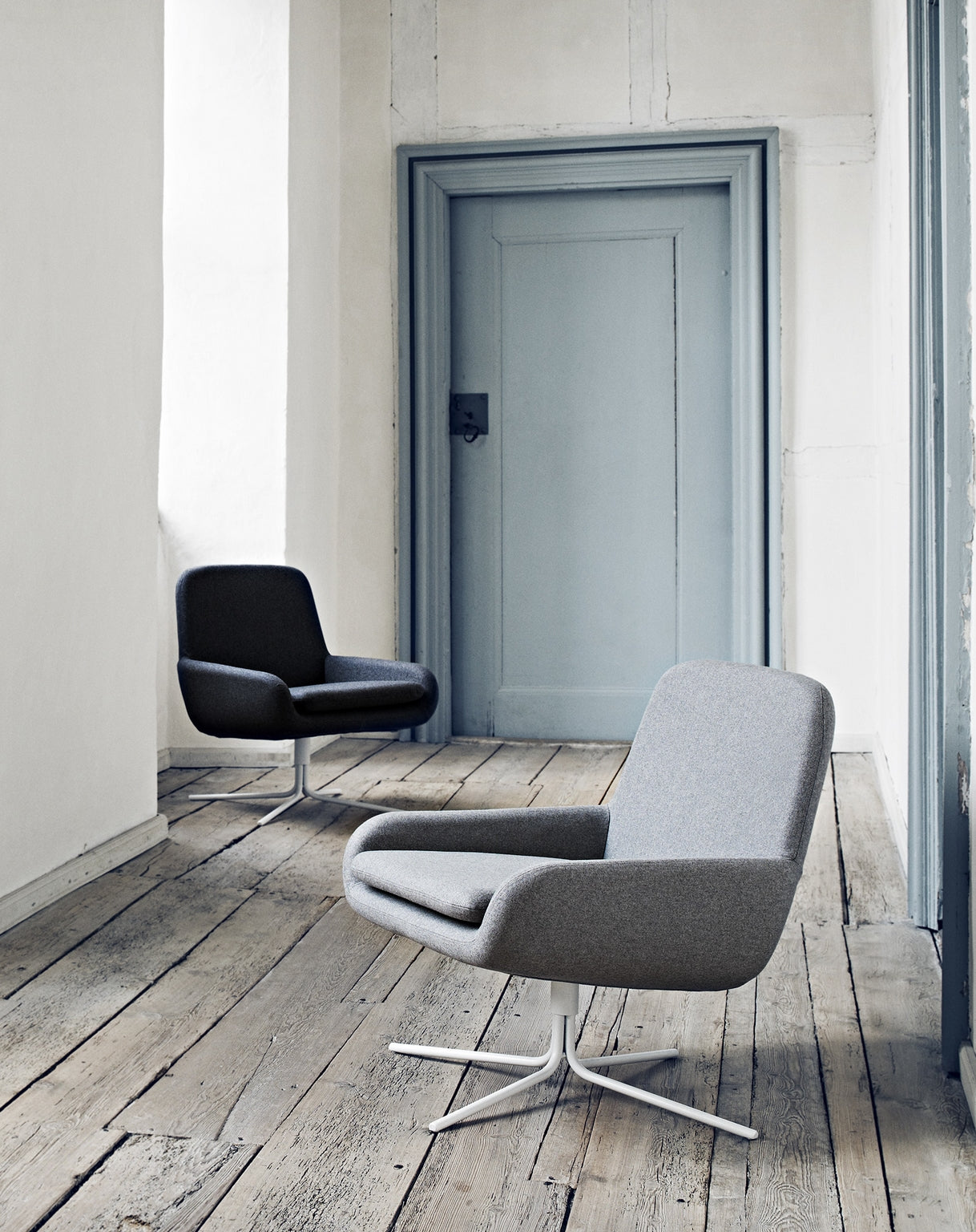 Deux chaises Fauteuil Pivotant Boreale de Busk+Hertzog pour Monsieur Bureau, en gris moderne avec des bases pivotantes en métal, reposent sur un sol en bois rustique dans un couloir minimaliste avec des murs blancs et une porte grise.#finitions_gris