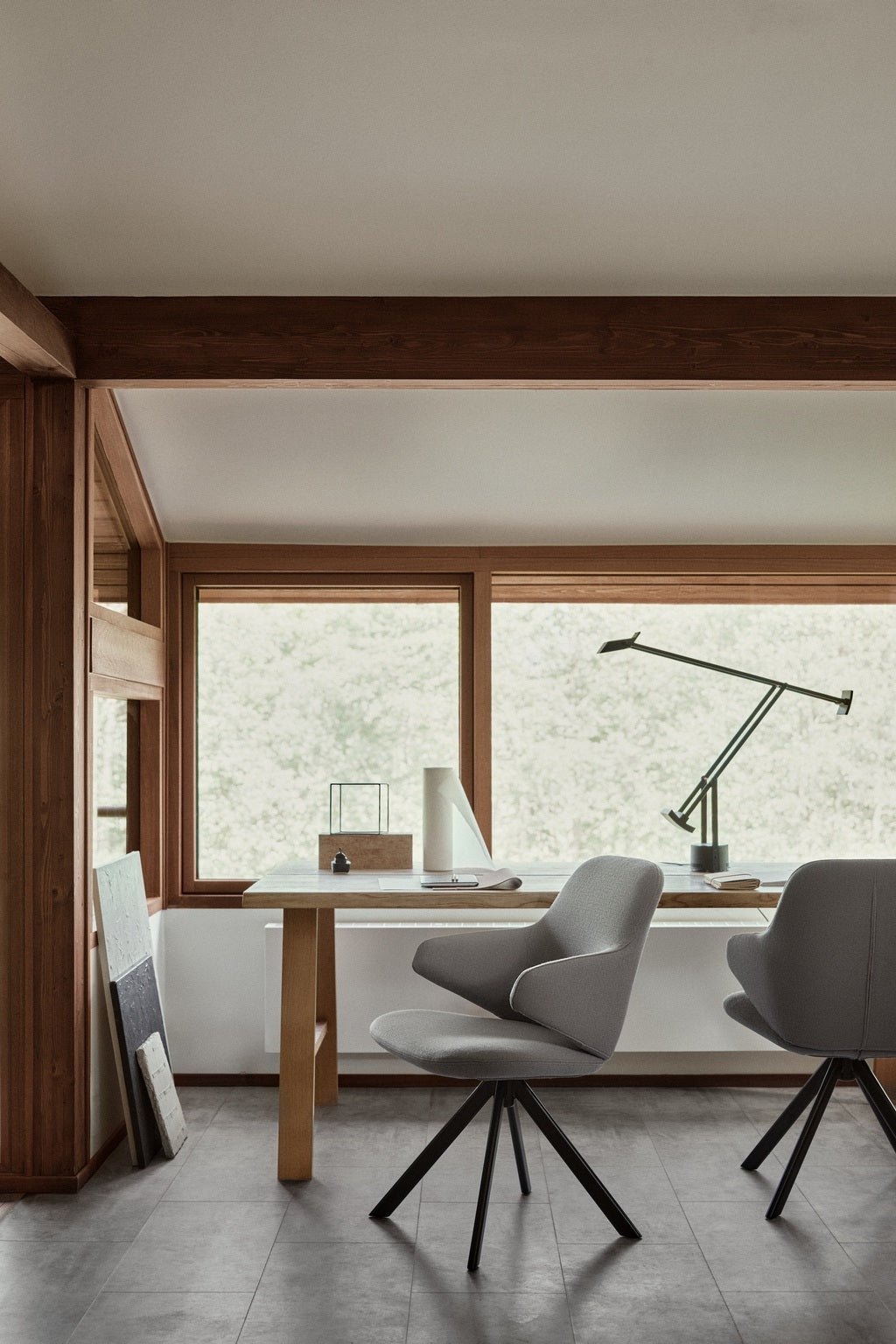 Bureau à domicile minimaliste avec de grandes fenêtres, deux chaises Monsieur Bureau Fauteuil pivotant design KAPP - Tissu Vision Gris, un bureau en bois, une lampe de bureau et des fournitures de bureau. La lumière naturelle met en valeur les lignes épurées et l'ambiance sereine de l'espace.