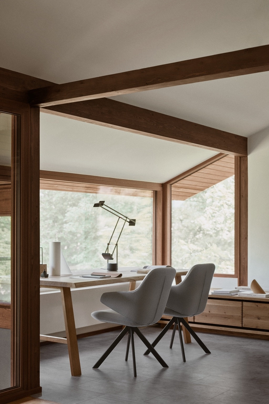 Un bureau à domicile moderne avec de grandes fenêtres, un bureau en bois, une lampe de bureau, des documents et deux fauteuils pivotants KAPP en tissu Vision Grey de Monsieur Bureau. Des poutres en bois et un décor minimaliste complètent l'espace, avec des arbres visibles à l'extérieur.