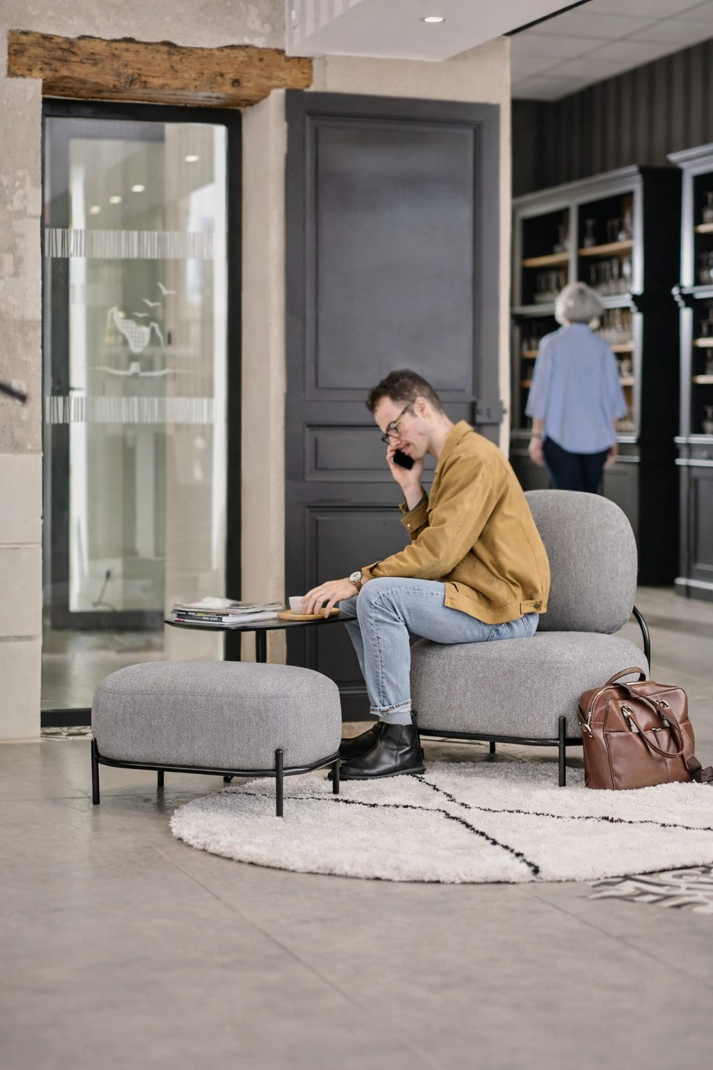 Un homme vêtu d'une veste brune est assis sur le Fauteuil d'accueil Brooklin - Pieds métal de Monsieur Bureau, en train de parler au téléphone et d'examiner des documents. Un sac brun est posé sur le sol, tandis qu'une chauffeuse d'accueil se fond dans le décor minimaliste.