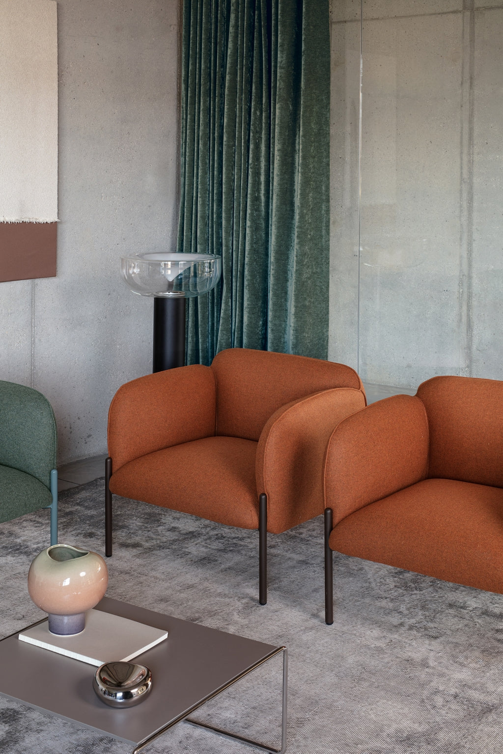 Un salon moderne avec deux fauteuils Fauteuil d'accueil design Kahlo de Monsieur Bureau, un rideau en velours vert, un bol en verre sur un support noir, un vase en céramique vert, un plat métallique sur une table basse grise sur un tapis gris texturé.