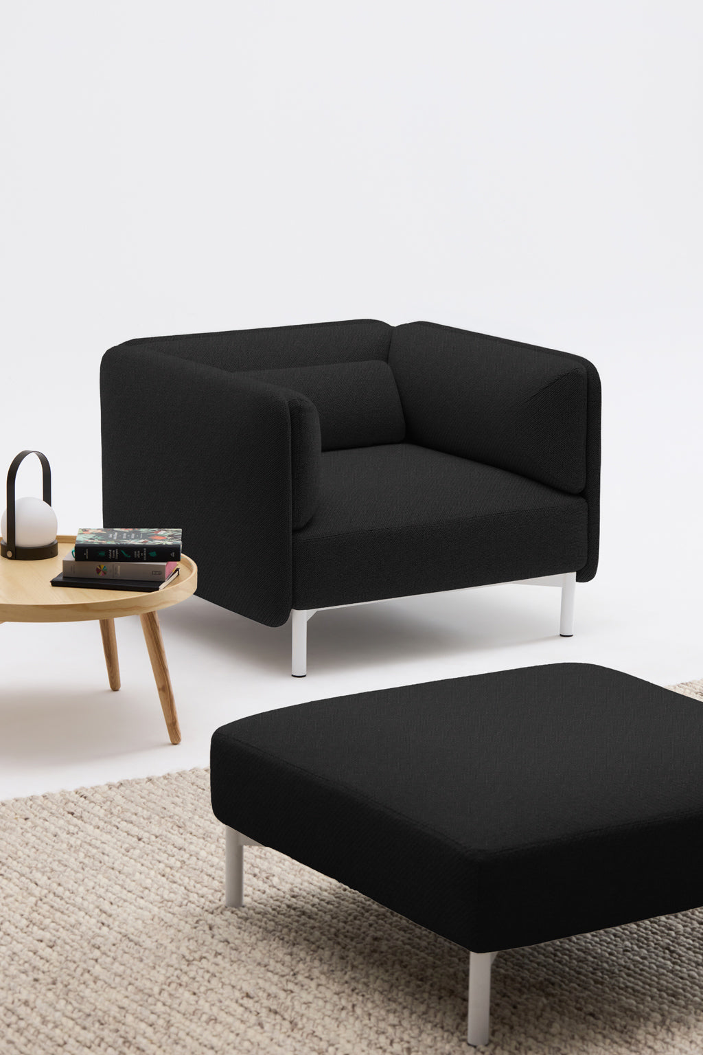 Un Fauteuil d'accueil modulaire Aurea de Monsieur Bureau en noir avec des pieds et un ottoman blancs est posé sur un tapis beige à côté d'une table d'appoint en bois clair avec des livres empilés et une lampe noire et blanche, le tout sur un fond blanc uni.
