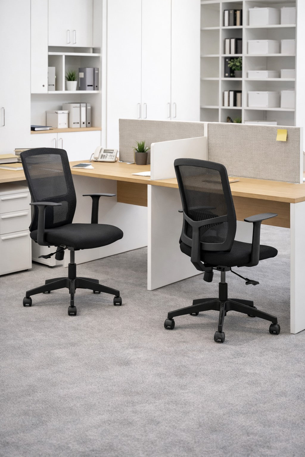 Deux chaises Monsieur Bureau Fauteuil de Bureau Mensa Basculant Centré en noir sont placées à un bureau commun en bois dans un bureau moderne et lumineux avec une moquette grise, des étagères blanches, des armoires, des plantes et des fournitures organisées en arrière-plan.