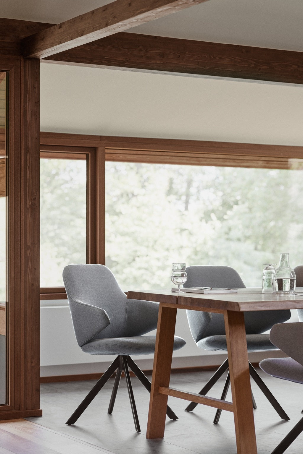 Un coin repas moderne avec une table en bois, des chaises Monsieur Bureau Fauteuil pivotant design KAPP - Tissu Vision Gris, et de grandes fenêtres qui laissent entrer la lumière naturelle. Des verres et une bouteille d'eau en verre sont posés sur la table, avec de la verdure visible à l'extérieur.