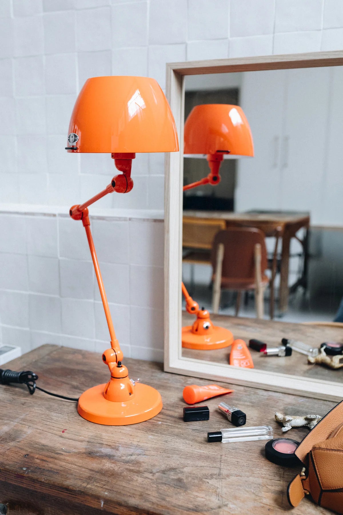 Une Lampe de bureau articulée Aicler design professionnel par Monsieur Bureau en orange vif est posée sur une surface en bois devant un miroir, avec des articles de maquillage et un sac de bronzage à proximité. La scène comprend des murs carrelés blancs et une chaise en bois.#finitions_orange