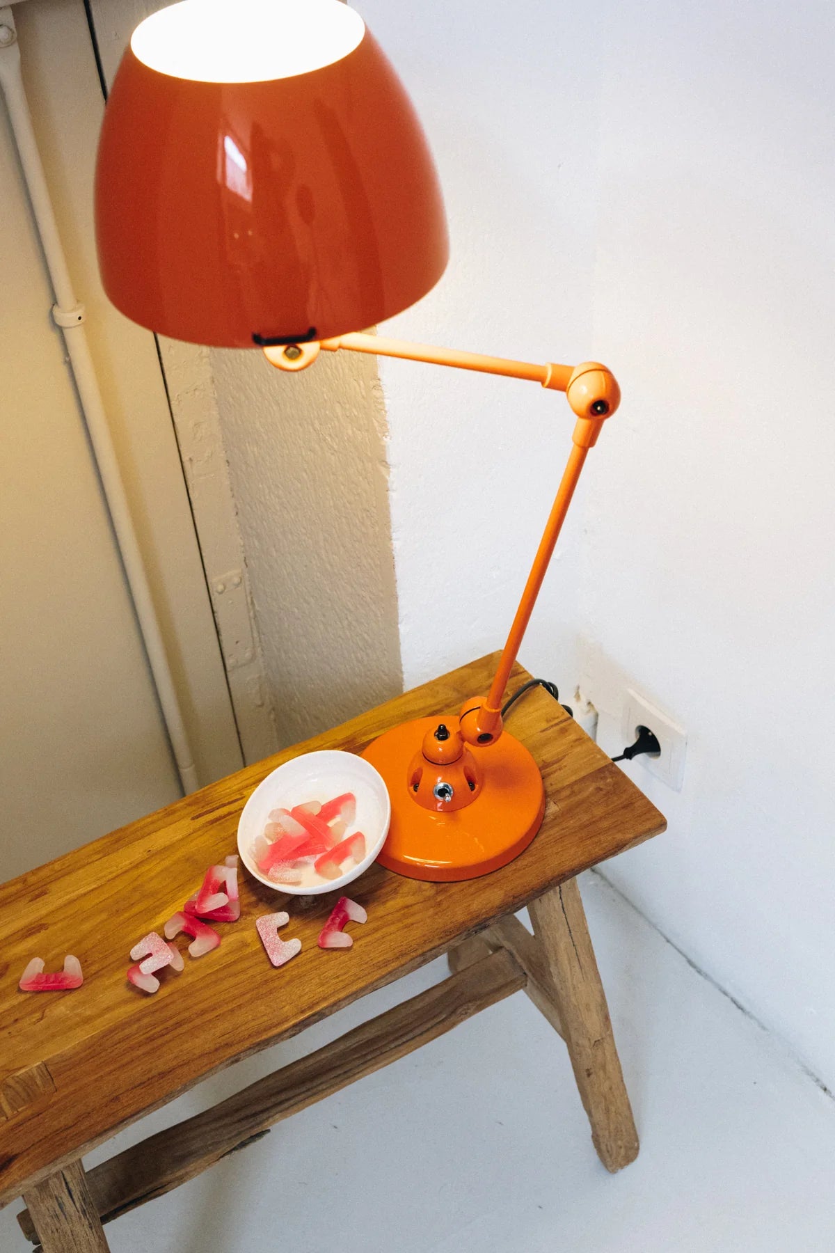 A Monsieur Bureau Lampe de bureau articulée Aicler design professionnel projette un éclairage directionnel sur un banc en bois avec un bol blanc de bonbons gélifiés roses et de sucreries éparses, branché sur une prise murale à proximité.