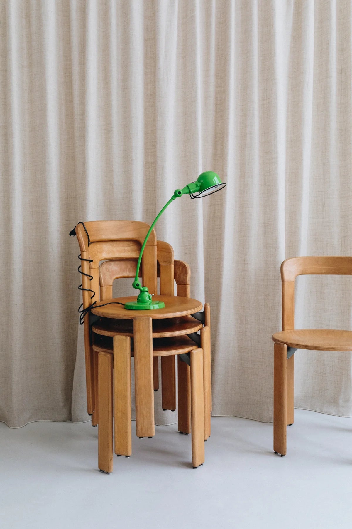 Une pile de trois chaises en bois se trouve devant des rideaux beiges, surmontés d'une Lampe de bureau articulée courbée Signal design de Monsieur Bureau, d'un vert éclatant. Une autre chaise en bois assortie se trouve à droite.#finitions_vert