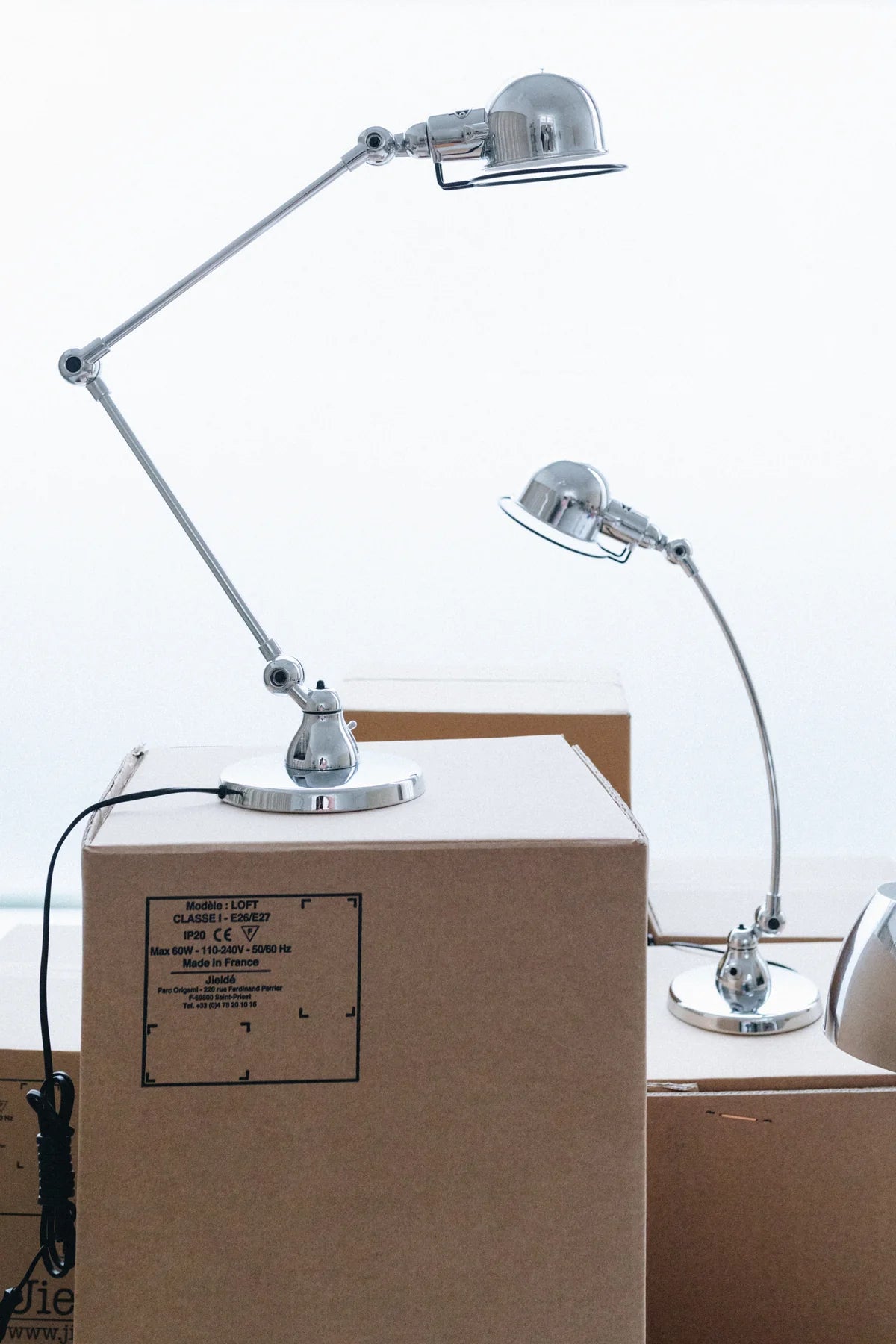 Deux Lampe de bureau articulée design Signal de Monsieur Bureau sont posées sur de grandes boîtes en carton dans une pièce lumineuse, évoquant une scène en mouvement. L'une est placée sur une boîte haute, l'autre sur une boîte plus basse, toutes deux fournissant un éclairage professionnel flexible.
