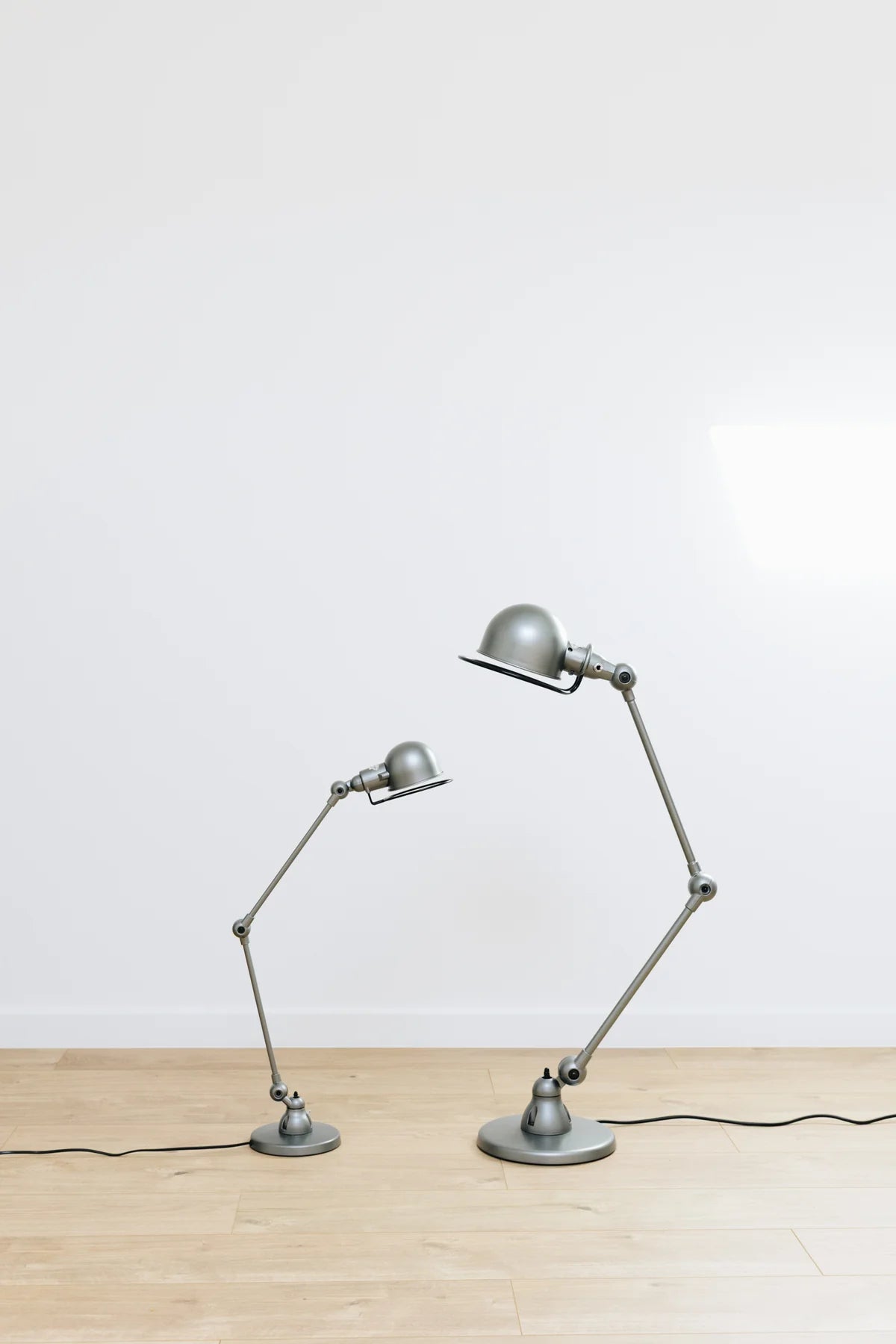 Deux lampes de bureau articulées design Signal de Monsieur Bureau, une petite et une grande, reposent non branchées sur un sol en bois clair devant un mur blanc. Idéales pour l'éclairage professionnel.