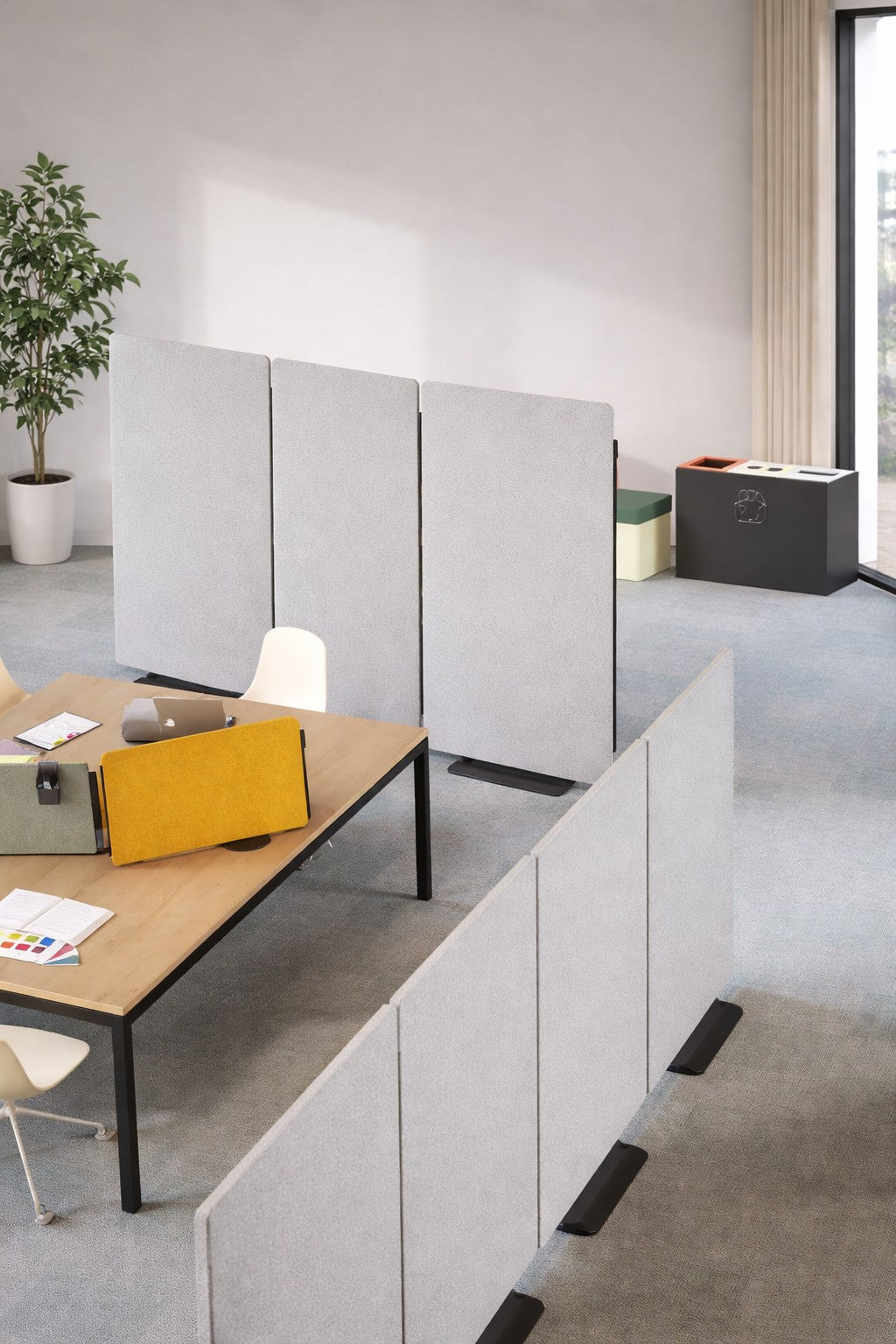 Bureau moderne en open space avec Monsieur Bureau Cloison Acoustique Silenza H160 avec Zip, un bureau en bois avec des ordinateurs portables, un écran jaune, des documents, des chaises blanches, une plante en pot, et des boîtes de rangement près d'une grande fenêtre pour le confort et la lumière naturelle.