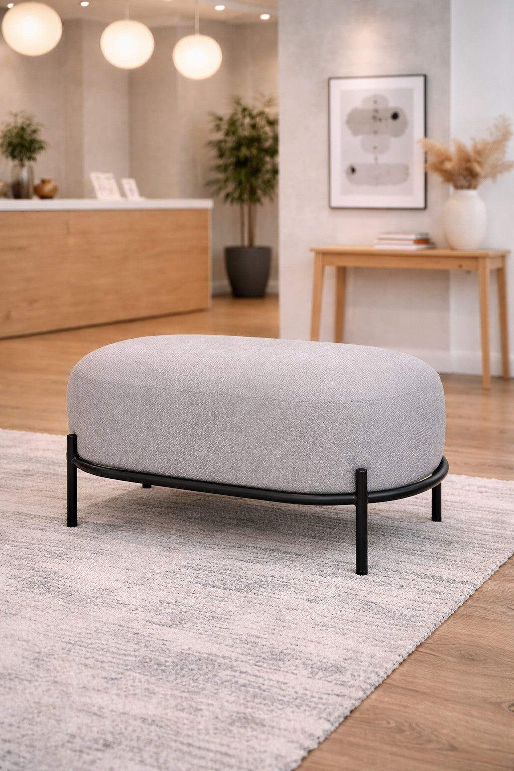 Le Pouf d'accueil Brooklin - Pieds métal de Monsieur Bureau, un pouf moderne ovale gris avec des pieds en métal noir, repose sur un tapis texturé dans une pièce minimaliste avec des planchers en bois et un éclairage doux - idéal comme assise d'appoint.