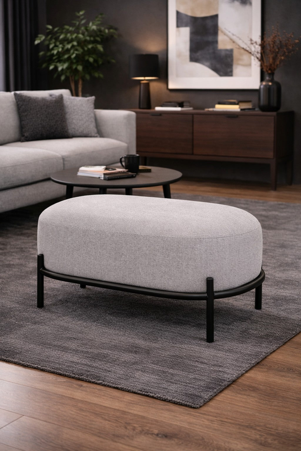 Un salon moderne avec le Monsieur Bureau Pouf d'accueil Brooklin - Pieds métal en gris clair sur un tapis foncé, une table basse ronde noire, un canapé gris, un buffet en bois et une décoration contemporaine avec une œuvre d'art abstraite et une plante en pot.