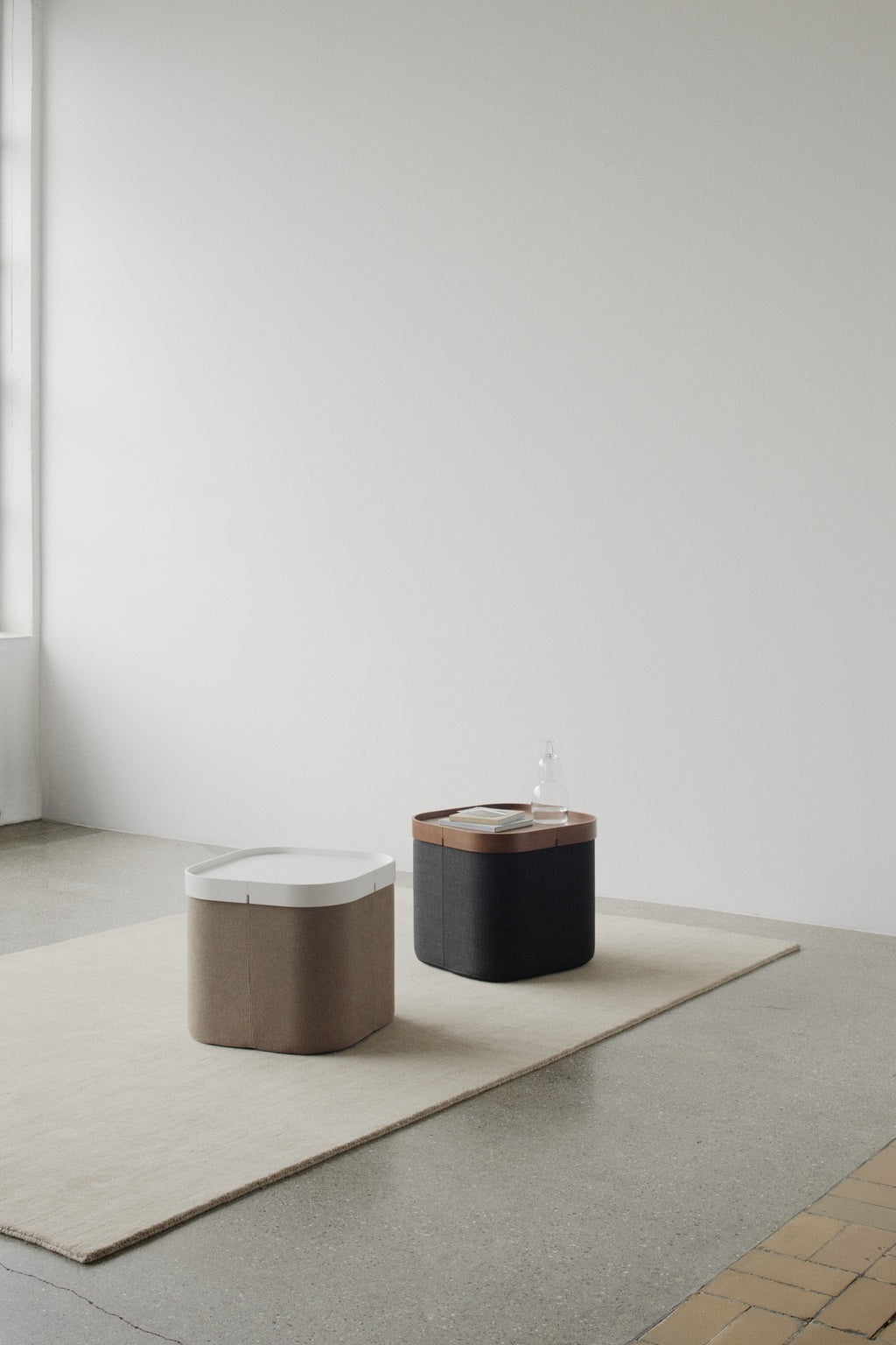 Dans une pièce minimaliste, deux tables d'appoint modernes en forme de boîte sont posées sur un tapis beige ; à proximité, un Pouf d'accueil Nordic avec plateau - Design Busk+Hertzog de Monsieur Bureau rehausse l'aspect neutre et épuré de la pièce.