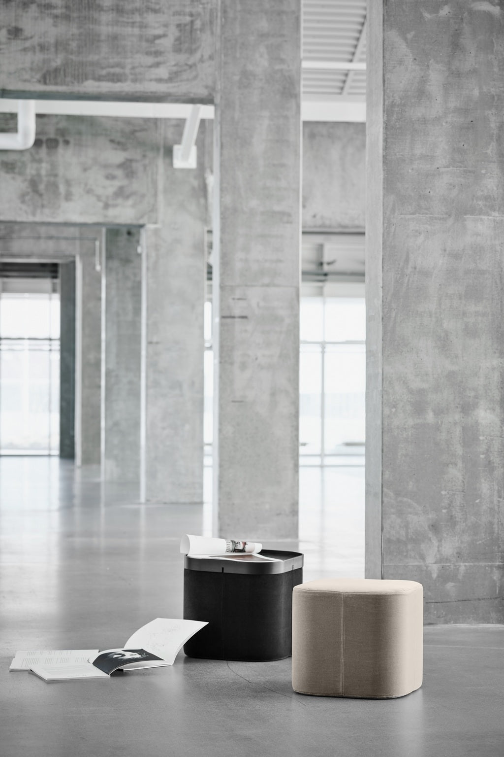 Un intérieur moderne et minimaliste avec des murs et des sols en béton présente le Pouf d'accueil Nordic avec plateau de Busk+Hertzog pour Monsieur Bureau, une boîte de rangement noire, des magazines ouverts et de grandes fenêtres qui inondent l'espace de lumière naturelle.