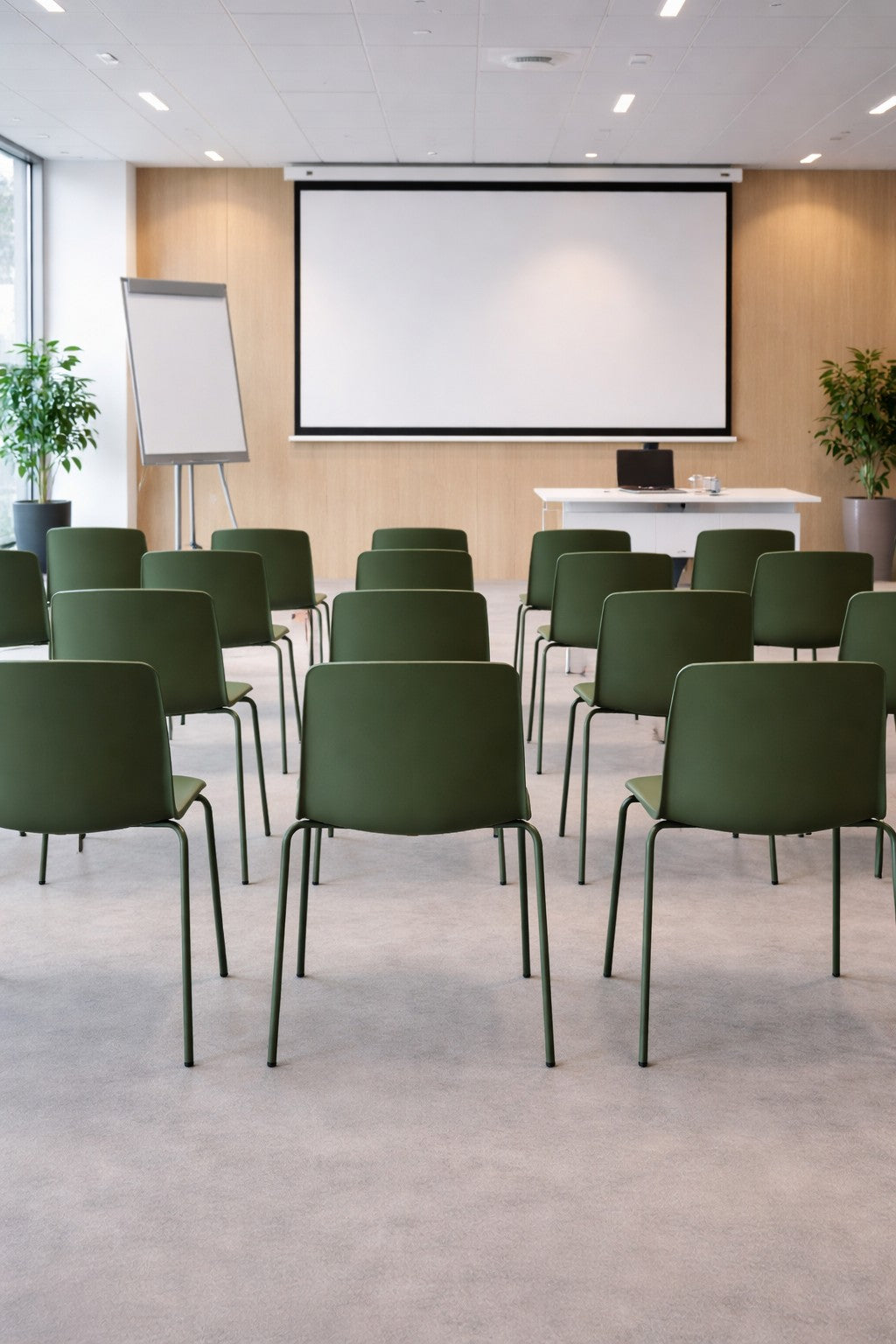 Des rangées de chaises empilables ALIX de Monsieur Bureau - un choix polyvalent, résistant et élégant - font face à un écran blanc et à un tableau à feuilles mobiles dans une salle de conférence moderne et lumineuse avec de grandes fenêtres et des plantes en pot.