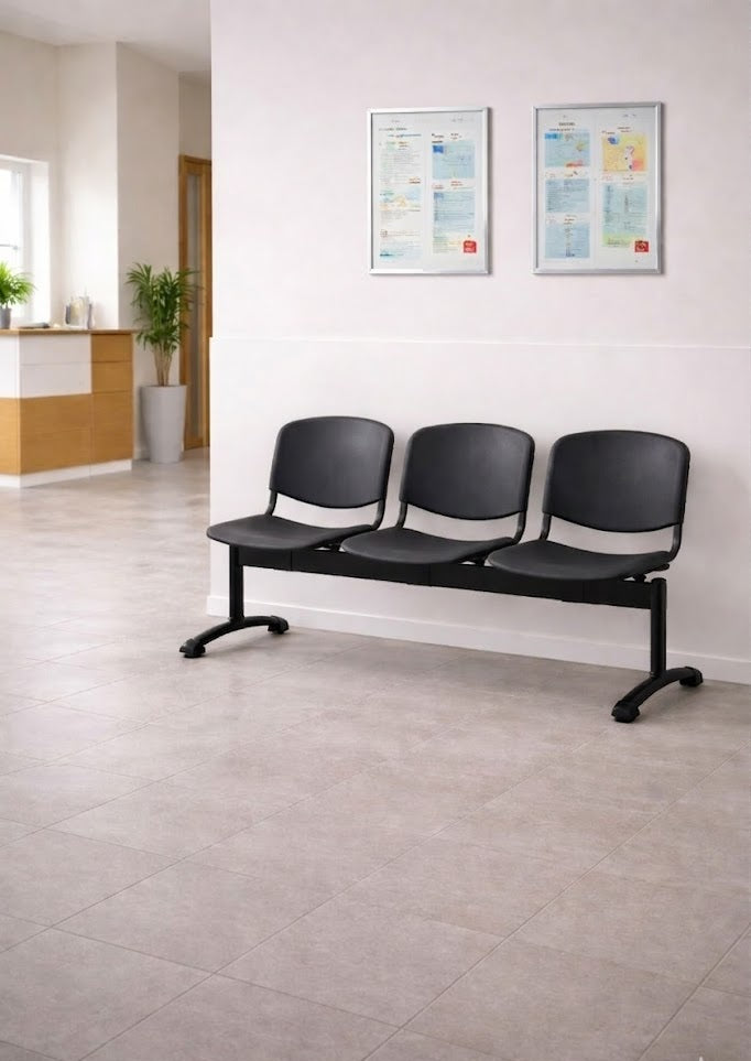 Trois places noires vides Sièges sur Poutre INO-P 3 de Monsieur Bureau sont alignées contre un mur clair, avec deux documents encadrés au-dessus. L'espace, propre et lumineux, est carrelé et une plante se trouve à l'arrière-plan, près du bureau d'accueil.#combo_3_places_noir
