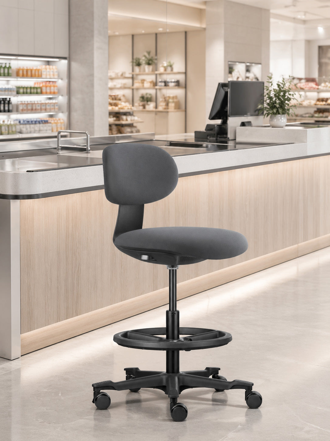 Le Siège technique Flex avec repose pieds de Monsieur Bureau, doté d'un repose-pieds circulaire et de roulettes, se trouve devant un comptoir aux lignes épurées, idéal pour le télétravail dans un magasin ou un café moderne et lumineux.