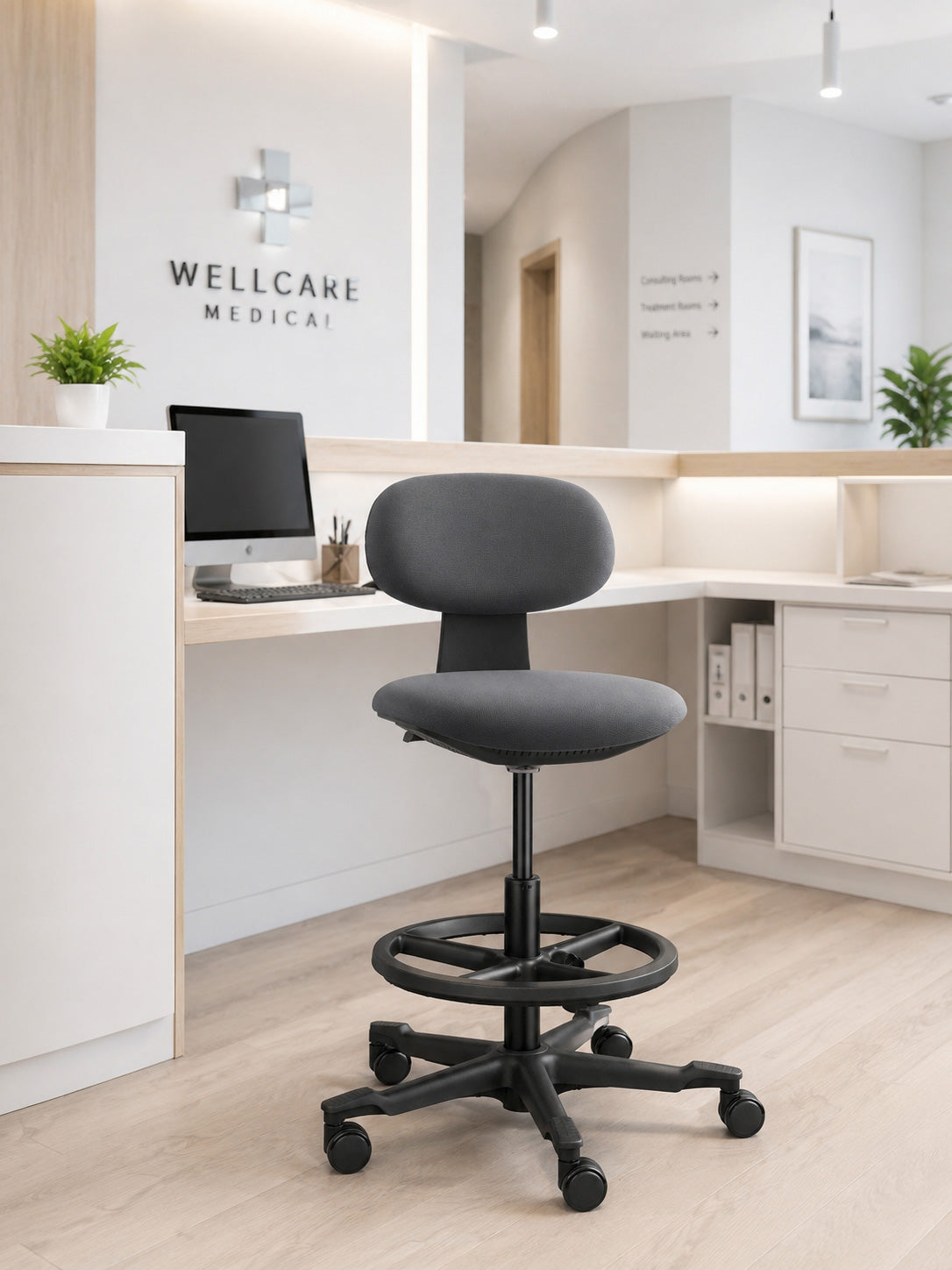 Un siège technique Flex avec repose pieds de Monsieur Bureau, sans accoudoirs, se trouve au centre de la réception d'un cabinet médical moderne avec un bureau, un ordinateur, des plantes, un meuble blanc et une enseigne Wellcare Medical sur le mur.