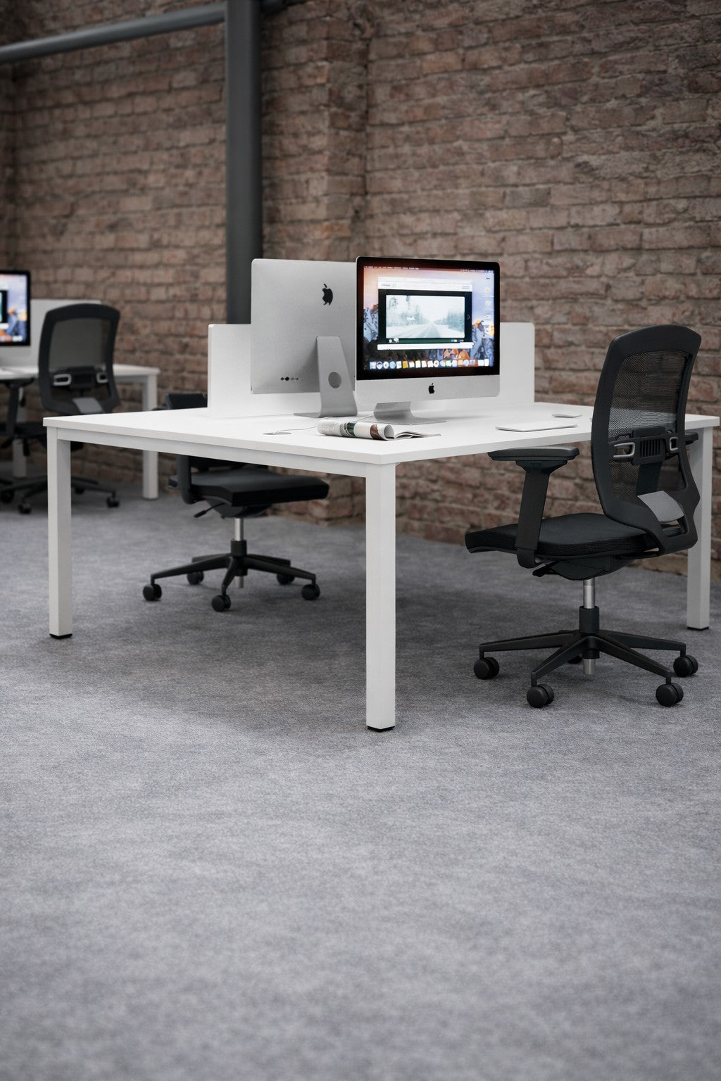 Un bureau moderne en open space avec des bureaux blancs Bureau Bench Astral pour 2 personnes P70 P80 cm de Monsieur Bureau, des chaises en maille noire, des iMacs, un sol en moquette grise et des murs en briques apparentes créant une atmosphère épurée et minimaliste.#combo_blanc_blanc