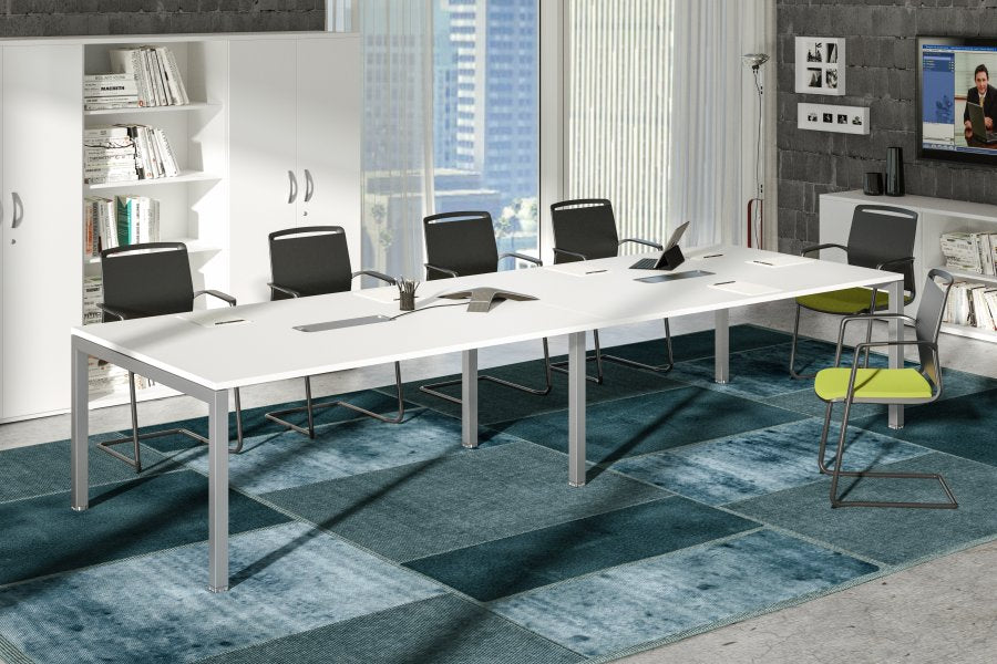 Salle de conférence moderne équipée du Monsieur Bureau Solution de Table 6 pers & Extensions Express+, de cinq chaises noires, d'une chaise verte, d'étagères avec des livres et d'une vue sur la ville à travers de grandes fenêtres. Un écran de télévision est fixé au mur.