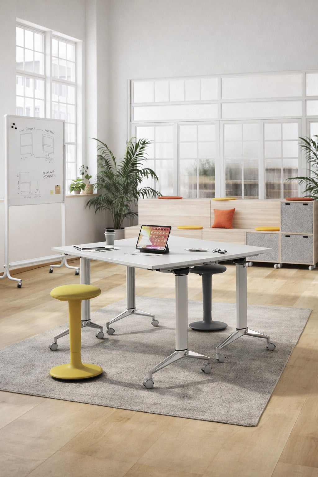 Bureau moderne comprenant une Table abattante Mobile sur roulettes de Monsieur Bureau, deux tabourets ergonomiques, un ordinateur portable, une tasse de café, des plantes, un tableau blanc et un banc rembourré sous de grandes fenêtres avec lumière naturelle.#combo_blanc_blanc