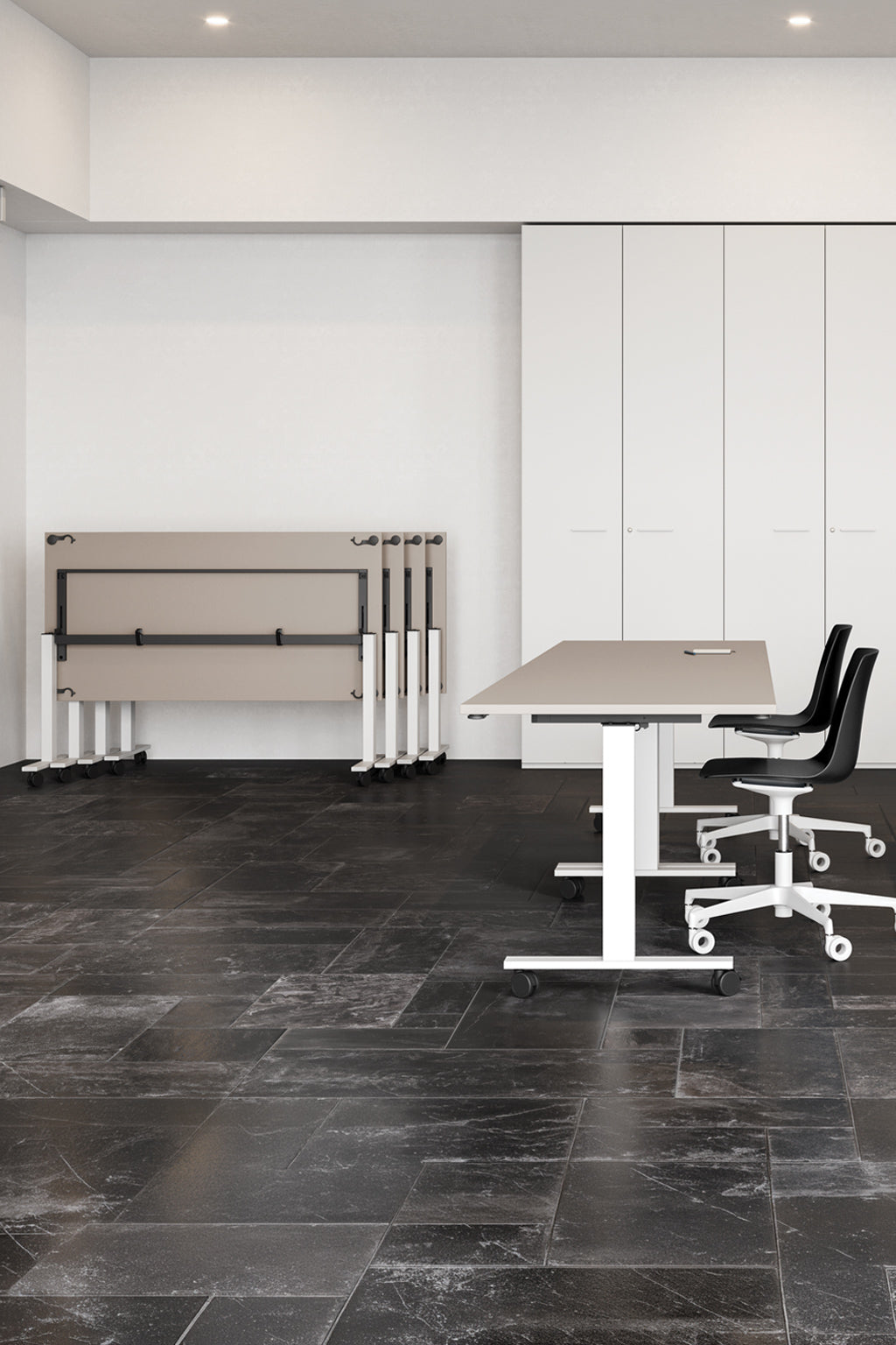 Bureau minimaliste avec des chaises pivotantes noires et des bureaux à roulettes sur des carreaux sombres ; un ensemble de Monsieur Bureau Table abattante Pop Folding sur roulettes (Profondeur 80 cm) est empilé le long d'un mur blanc avec des armoires de rangement encastrées à l'arrière-plan.