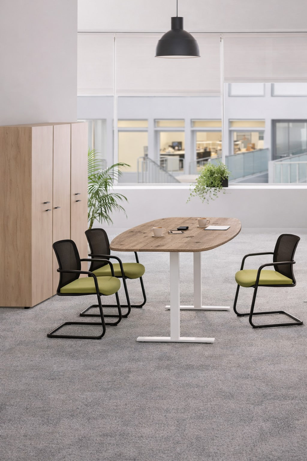 Un espace de réunion moderne avec la Table tonneau électrique Top & Down (6/8 personnes) de Monsieur Bureau, trois chaises noires avec des coussins verts, un meuble en bois, une plante, une lampe noire suspendue et de grandes fenêtres pour la lumière naturelle..#combo_timber_blanc