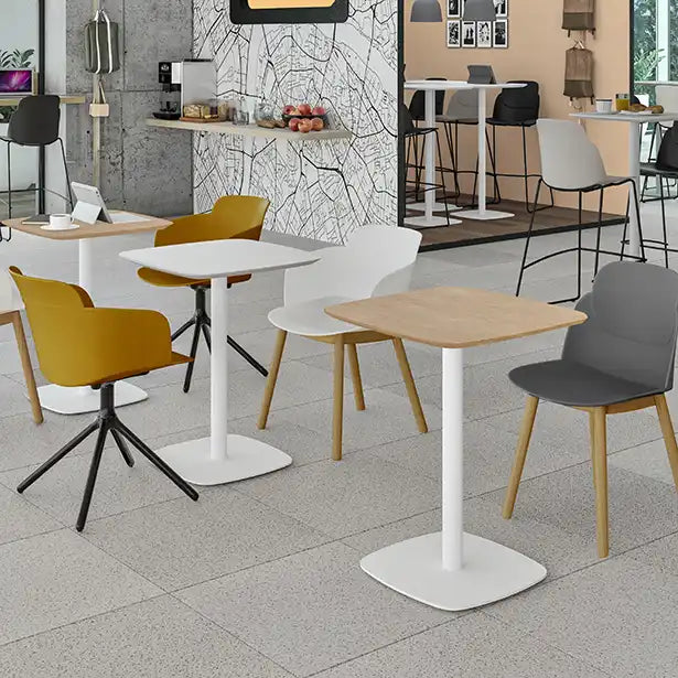 Un intérieur de café moderne avec un sol en carrelage blanc, une table de collectivité Kafeta de Monsieur Bureau (plateau en MDF) et des chaises jaunes, grises et blanches mélangées. À l'arrière-plan : un mur sur le thème de la carte, un petit comptoir et des sièges de bar.