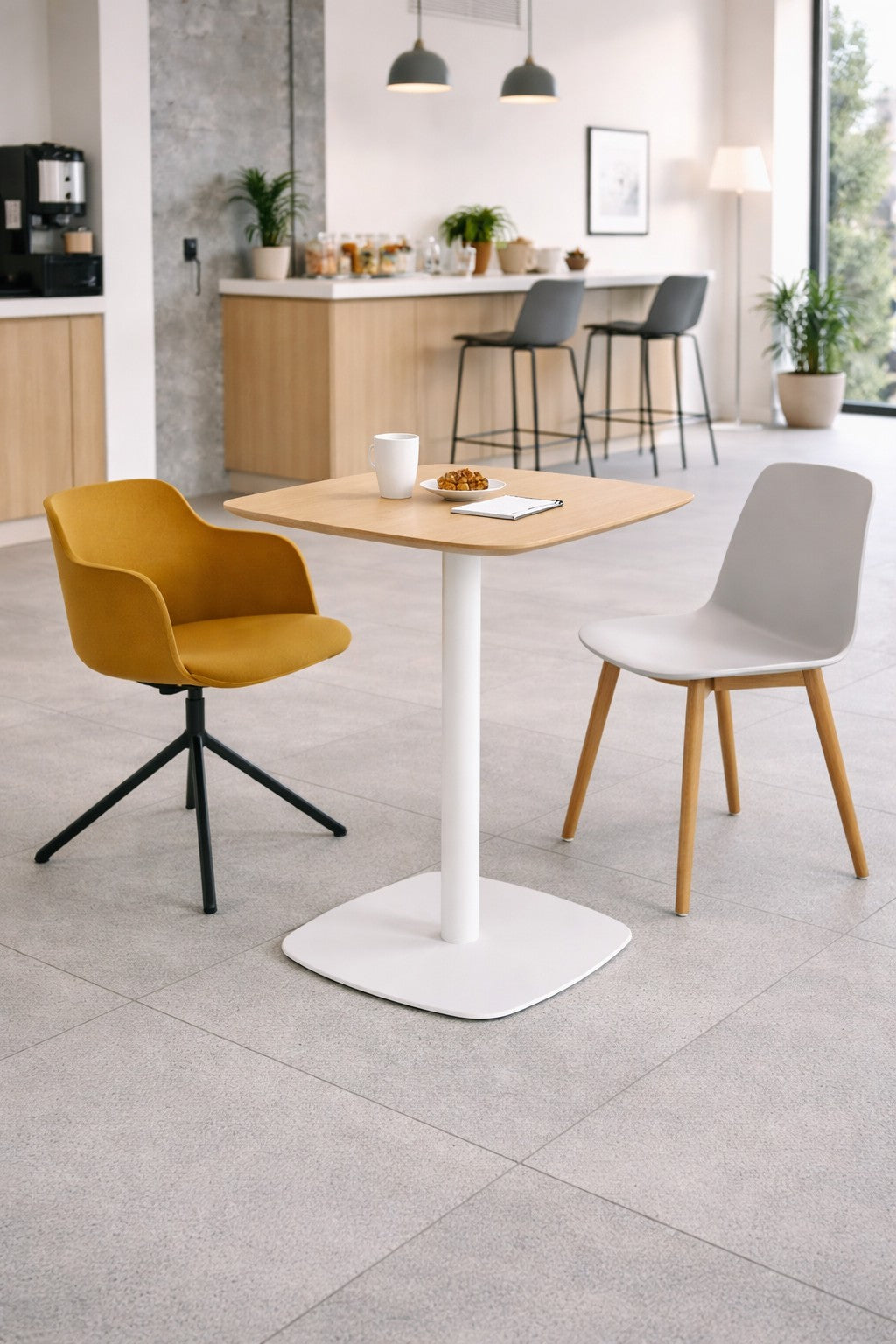 Un café moderne avec un Monsieur Bureau Table de collectivité Kafeta (plateau MDF), une chaise pivotante jaune et une chaise blanche. Sur la table : tasse blanche, biscuits, carnet de notes. En arrière-plan : comptoir, tabourets, plantes, grandes fenêtres.