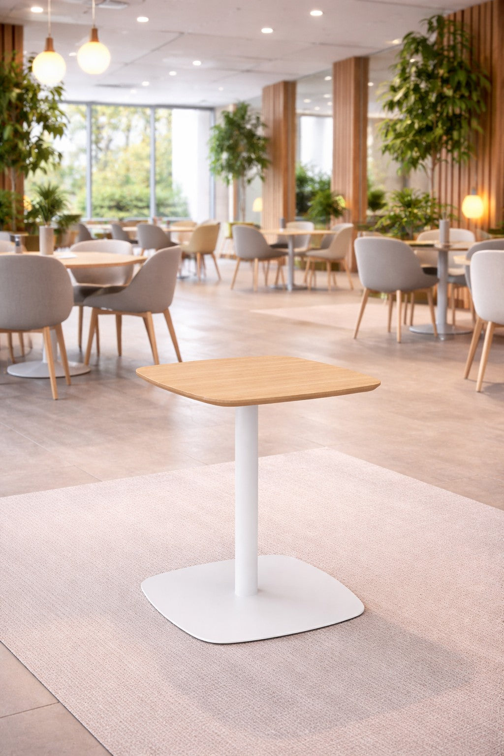 Un café moderne avec des meubles en bois clair, des chaises blanches et de grandes fenêtres. Un Monsieur Bureau Table de collectivité Kafeta avec un plateau en MDF et une base blanche est centré sur un tapis clair au premier plan parmi des plantes d'intérieur.#finitions_bois
