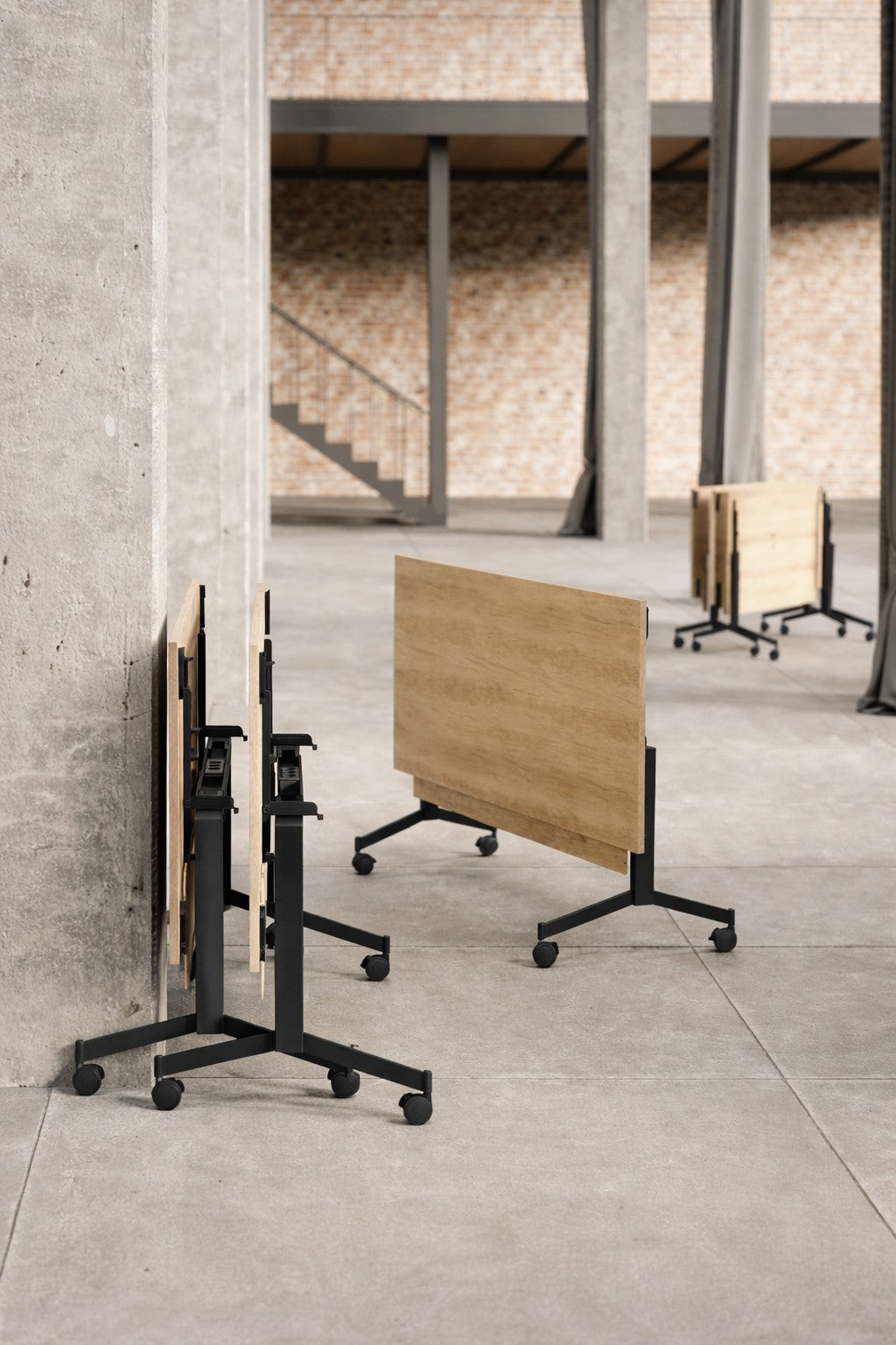 Plusieurs tables Réunion Abattante et Mobile Quick Moove de Monsieur Bureau, avec des plateaux en bois et des cadres en métal noir, sont pliées et disposées sur un sol en béton dans un espace industriel ouvert avec des murs en briques et de hautes colonnes.#combo_chene_nebrasa_noir