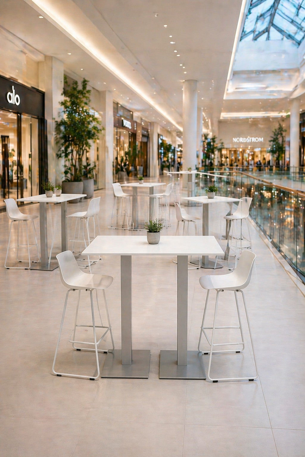 Un centre commercial intérieur moderne où l'on peut s'asseoir, avec une Table haute Newline rectangulaire - Pieds carrés de Monsieur Bureau, des plantes en pot sur chaque table, et des magasins comme alo et Nordstrom en arrière-plan, éclairés par la lumière naturelle.