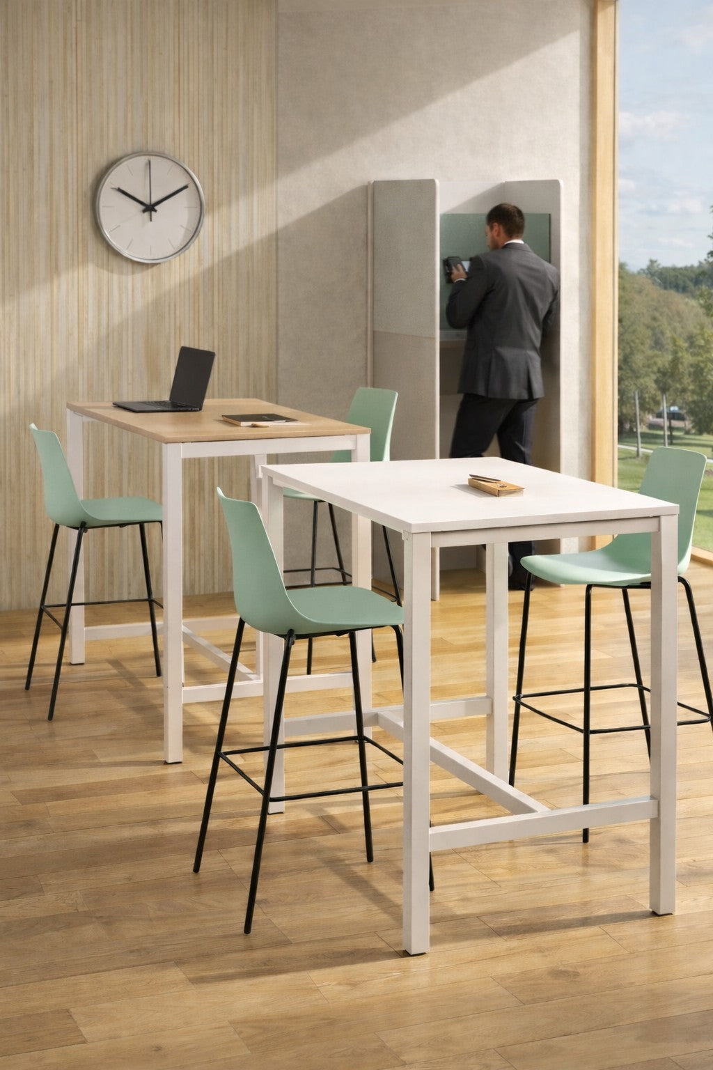 Un bureau moderne est équipé de deux tables Haute Astral 4 à 8 Pers 120 à 160 cm de Monsieur Bureau, de chaises vertes, d'une horloge murale, d'un ordinateur portable et d'un carnet de notes. Une personne en costume se tient à une cabine murale près d'une grande fenêtre avec vue sur la verdure.