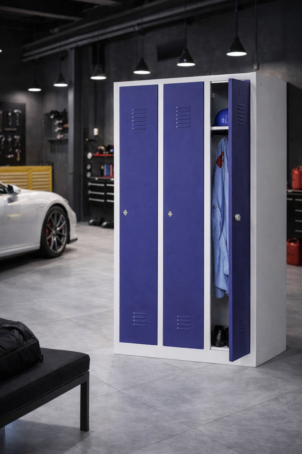 Un Vestiaire métallique Talos 1 porte - 1 à 3 colonnes bleu et blanc de Monsieur Bureau est ouvert dans un garage moderne, présentant un casque, une combinaison bleue et des chaussures, avec une voiture blanche et des établis noirs à proximité dans l'espace industriel.